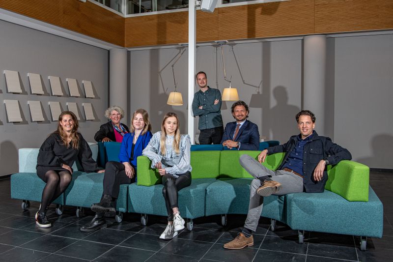 Op deze foto zie je een groep mensen afgebeeld. De meeste mensen zitten op een groen/grijze bank in een grote ruimte. Van links naar rechts zie je op de foto: Eva Legemate, Ilona Vermare Hoefkens, Rosanne Houterman, Noami Braat, Pieter-Bart Visscher en Jeroen Vreeke. Pim van Liere staat achter de bank, linksachter Pieter-Bart